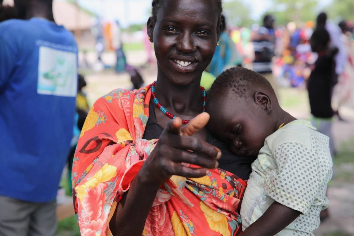 En kvinne peker på kamera og holder sin sovende baby mens familier står i kø for å bli registrert for hjelp og matutdeling i Sør-Sudan. Foto: UNICEF/UN0230481/Elrington.